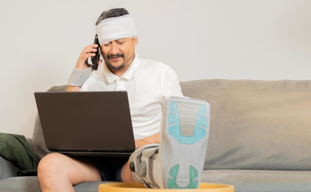 A man with bandages on his head and a cast on his foot, speaking to someone on his phone while typing on a laptop
